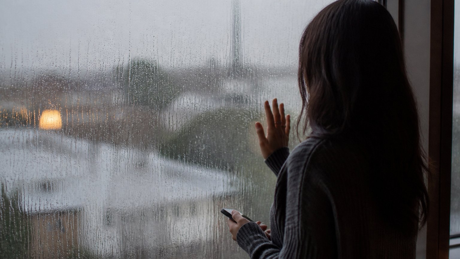 A person gazing out a rain-streaked window lost in thought, preoccupied with a relationship
